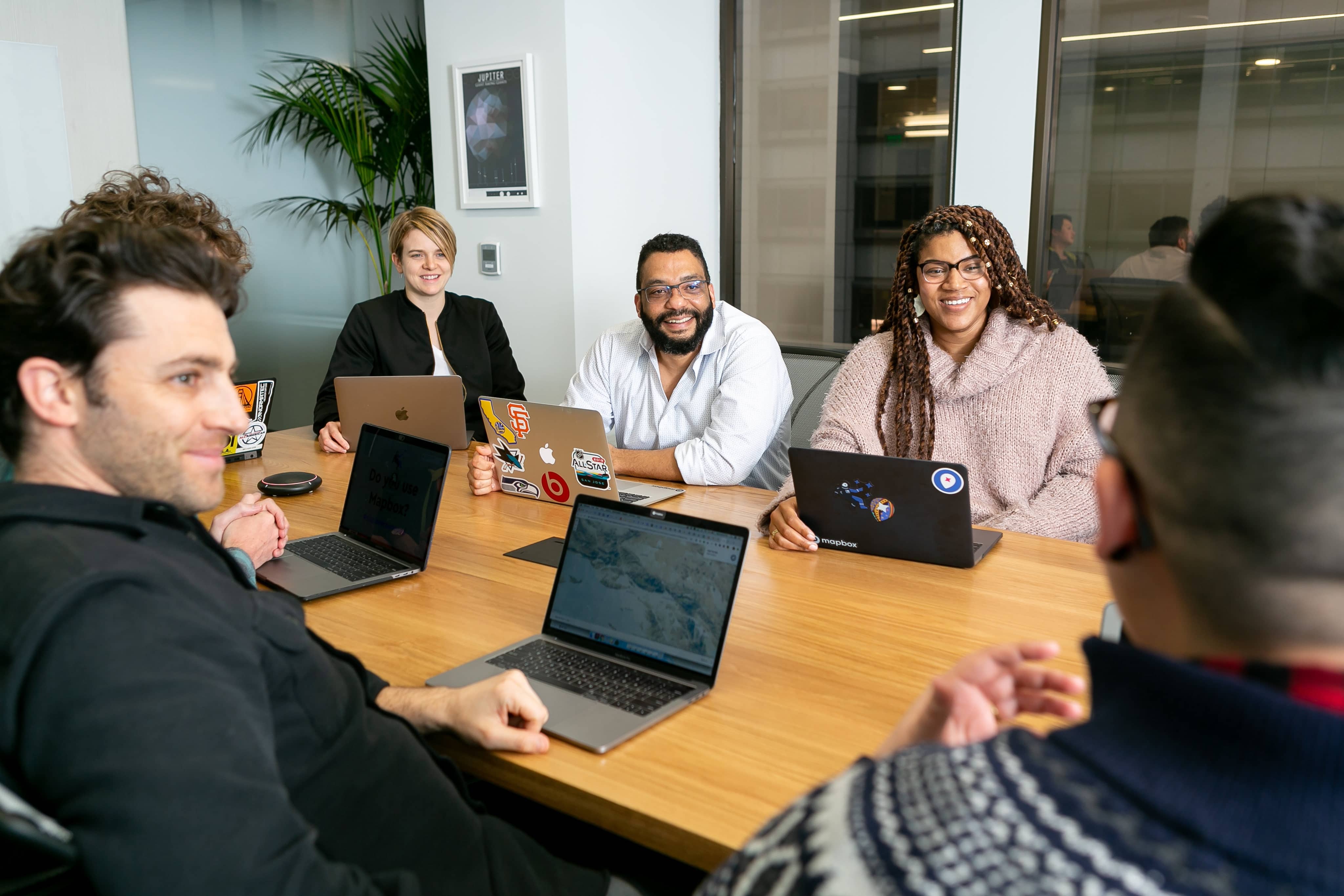 People collaborate at a desk with their laptops, listening to a colleague talking.