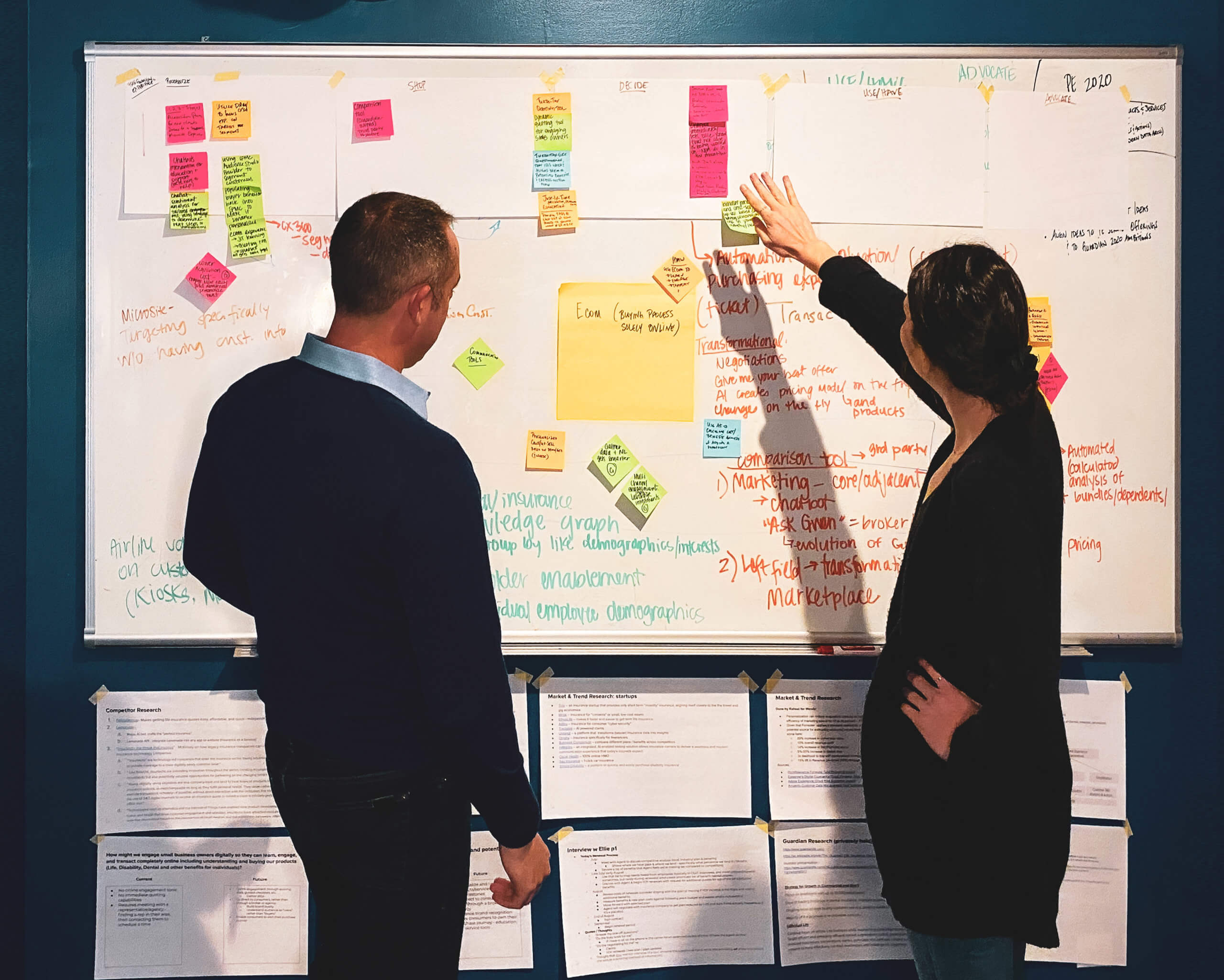 Two people look at a whiteboard with stickies and workshop notes.