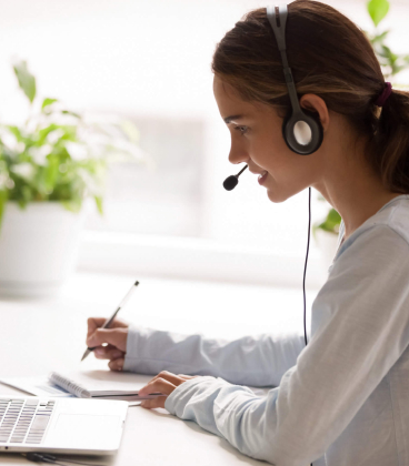 A person with a headset writes notes while looking at a laptop.