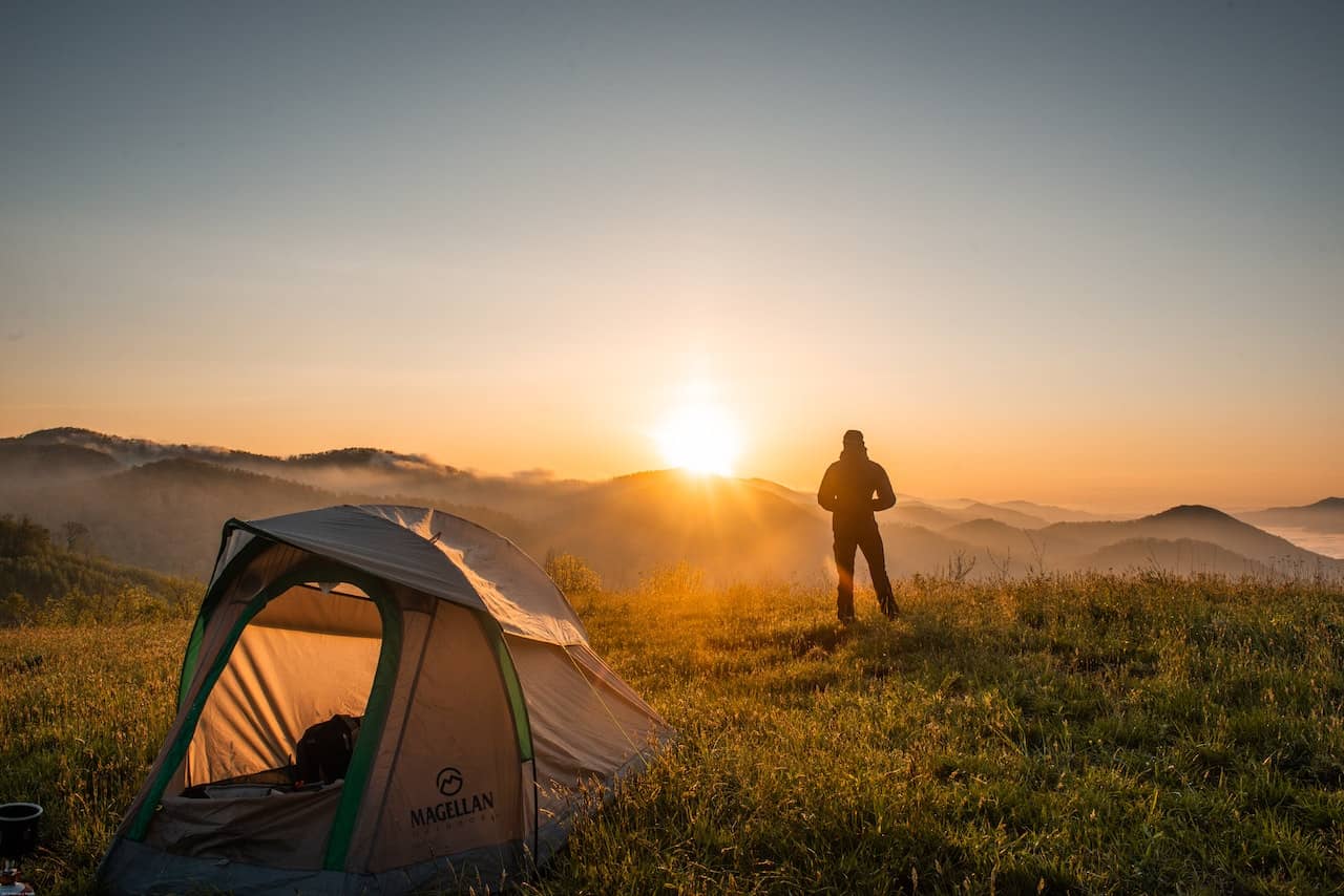 A man standing in front of a sunrise with a tent nearby in nature.