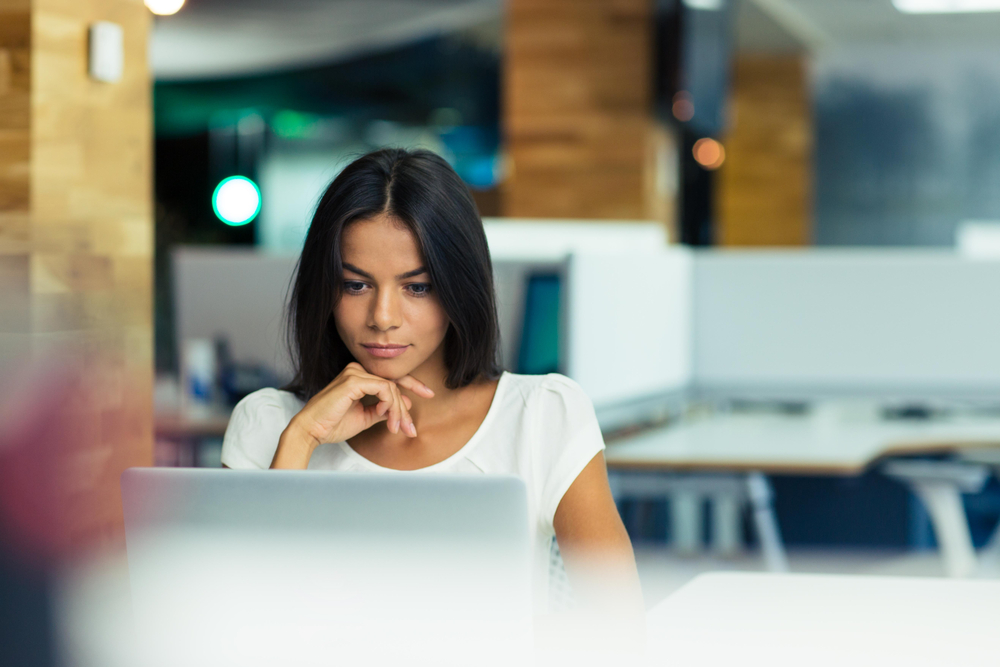 A woman rests her chin on her hand, looking thoughtful while looking at a computer screen.