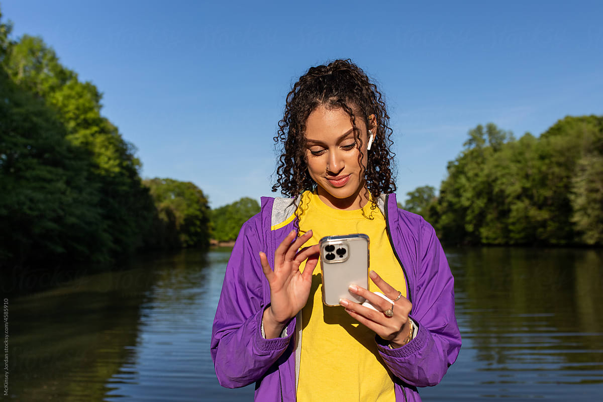 A person wearing earbuds touches their phone with a lake behind them.